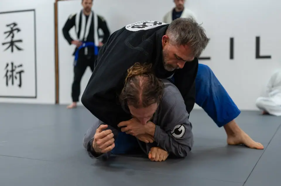 Two grapplers in black and blue gis working from the mat at Ascended BJJ, jiu-jitsu characters on the wall behind them