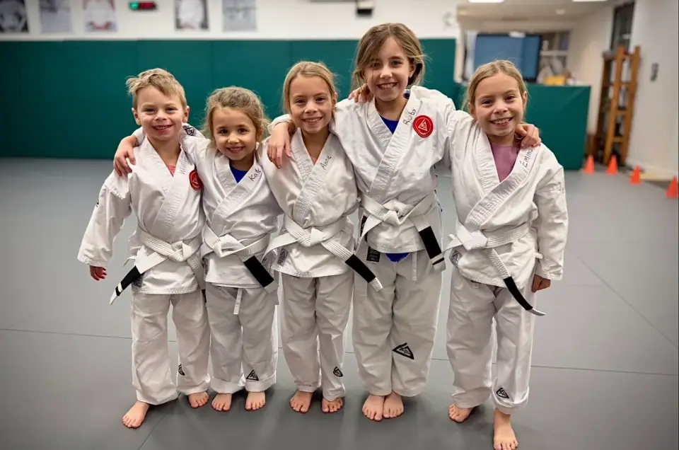 Five smiling girls in white gis standing arm in arm on the mat at Gracie Jiu Jitsu St. Nicolas