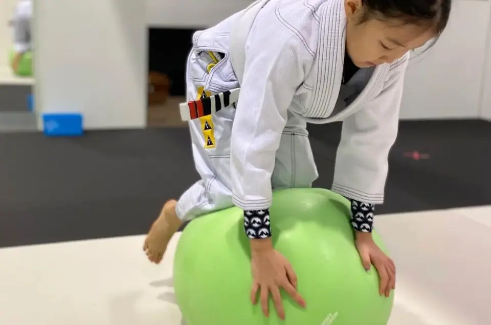A young student in a white gi climbing over a green exercise ball during a True North Jiu-Jitsu kids class